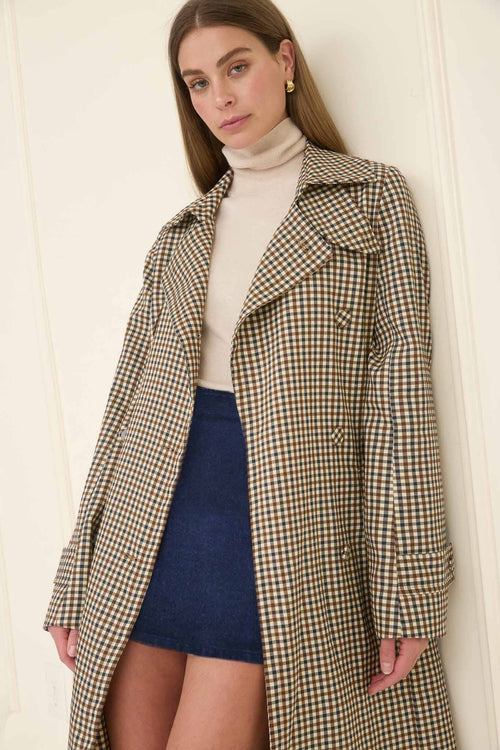 Woman wearing a checkered coat over a turtleneck and skirt against a white wall.