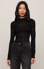 Woman wearing a black long-sleeve top with intricate patterns and high-waisted black jeans on a beige background