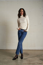 Woman wearing a cream turtleneck sweater and blue jeans standing against a plain white wall.