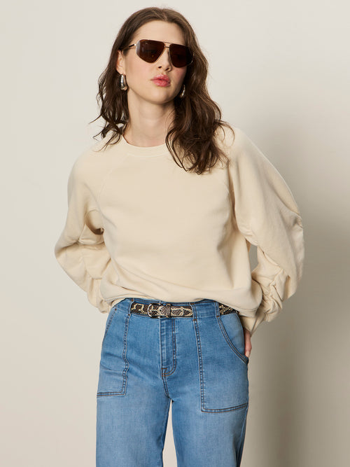 Woman wearing a beige sweater and blue jeans with sunglasses on a plain background