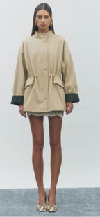 A model stands facing forward wearing a beige long-sleeve jacket with dark green plaid cuff accents, a high collar, and front flap pockets, paired with a short skirt and snakeskin heels against a light gray studio background.