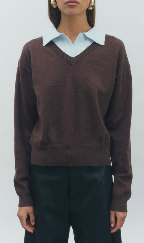 A woman wearing a dark brown v-neck knit sweater layered over a light blue collared shirt, paired with black trousers, against a plain light gray background.