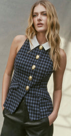 A woman wearing a sleeveless navy and black checkered vest with a white collar and gold buttons, paired with black leather pants, set against a neutral gray background.