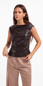 A woman modeling a dark brown, sleeveless draped top with a glossy finish, paired with beige pants, against a plain white background.