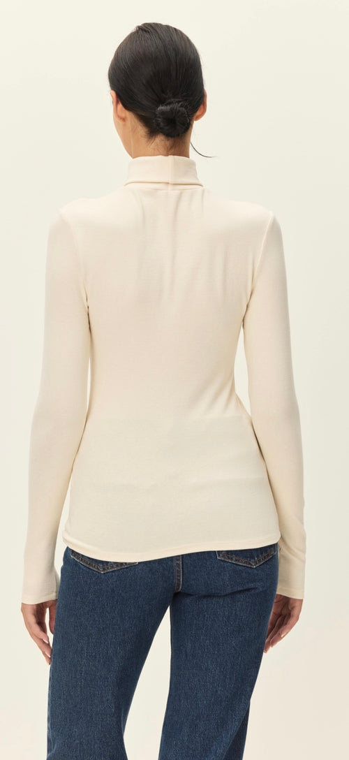 A woman shown from the back wearing a fitted long-sleeve mock-neck top in a light cream color, paired with dark blue jeans, against a plain off-white studio background.