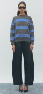 A model wearing a gray crewneck sweater with horizontal blue stripes, paired with wide-leg black trousers and black pointed shoes, standing against a light gray studio background.