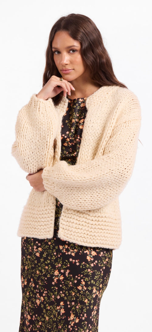 A woman modeling a chunky cream-colored knit cardigan over a black floral dress against a plain white background.