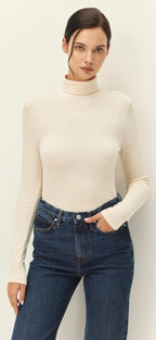 A woman modeling a fitted, long-sleeve cream mock neck top paired with dark blue jeans, set against a plain off-white studio background.