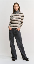 A model wearing a beige, black, and gray chevron-patterned knit mock-neck sweater paired with dark gray wide-leg jeans, standing against a light gray studio background.