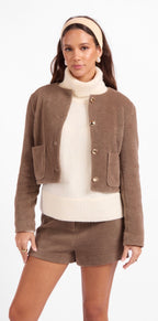 A woman modeling a brown textured cropped jacket with gold buttons over a cream turtleneck sweater and matching brown shorts, against a plain white studio background.