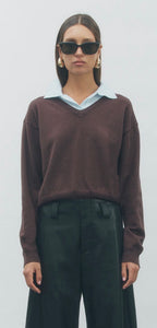 A model wearing a dark brown v-neck knit sweater with a light blue collar, paired with black wide-leg trousers and black sunglasses, against a plain light gray background.