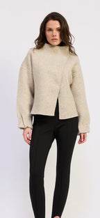 A woman modeling a light beige asymmetrical wrap-front cropped sweater with long sleeves, paired with black pants, against a plain white studio background.