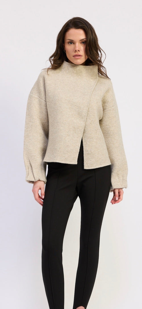 A woman modeling a light beige asymmetrical wrap-front cropped sweater with long sleeves, paired with black pants, against a plain white studio background.