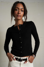 Woman wearing a black long-sleeve shirt and white pants against a plain background