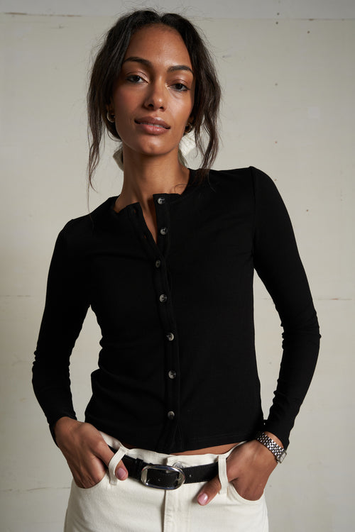 Woman wearing a black long-sleeve shirt and white pants against a plain background