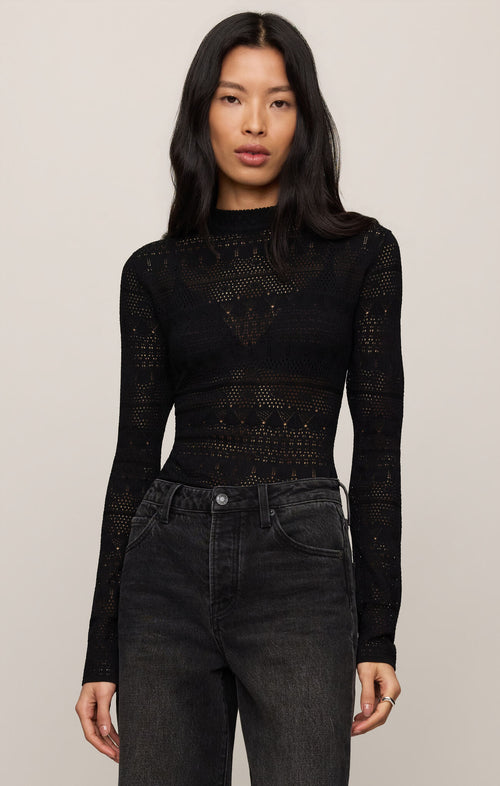 Woman wearing a black long-sleeve top with intricate patterns and high-waisted black jeans on a beige background