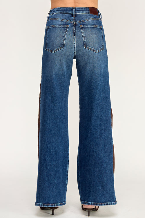 Rear view of a person wearing blue wide-leg denim jeans with dark brown side panels and black high heels, photographed against a plain light gray studio background.