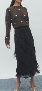 Woman wearing a brown sweater with white embellishments and a black fringed skirt on a light gray background