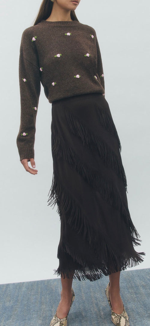 Woman wearing a brown sweater with white embellishments and a black fringed skirt on a light gray background