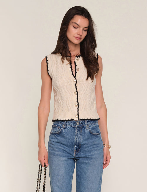 Woman wearing a sleeveless cream top with black trim and blue jeans on a plain background