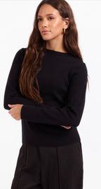 A woman modeling a black long-sleeve knit sweater with a wide boat neckline, paired with black trousers, against a plain white studio background.