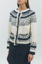 A woman wearing a cream and navy knit cardigan sweater with a Fair Isle pattern and brown buttons, paired with a dark skirt, against a light gray studio background.