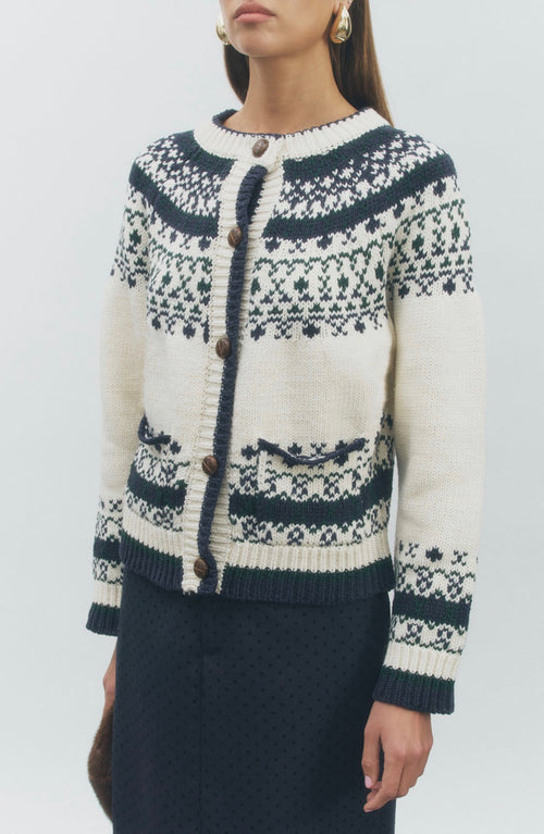 A woman wearing a cream and navy knit cardigan sweater with a Fair Isle pattern and brown buttons, paired with a dark skirt, against a light gray studio background.