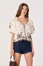 Woman wearing a cream blouse with black floral embroidery and denim shorts on a white background