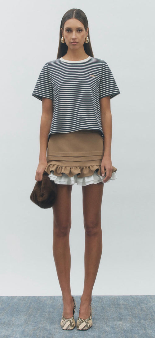 A model stands against a light gray studio background wearing a short-sleeve black and white striped t-shirt, a brown ruffled mini skirt with a white ruffle underneath, holding a dark brown handbag, and wearing snakeskin-patterned heels.