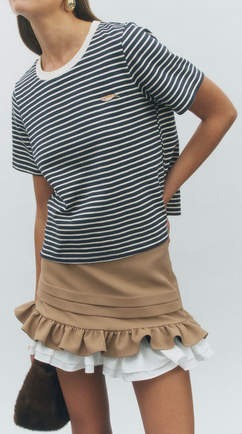 A woman wearing a short-sleeve black and white striped t-shirt with a small gold chest logo, paired with a tan ruffled mini skirt over a white ruffled underskirt, against a light gray background.