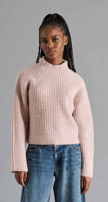 Person wearing a light pink ribbed sweater and blue jeans against a gray background