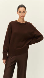A woman modeling an oversized dark brown knit sweater with ribbed cuffs and hem, paired with matching brown trousers, against a plain light beige studio background.