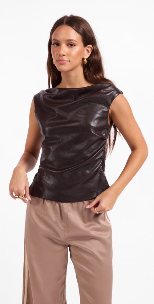 A woman modeling a dark brown, sleeveless draped top with a glossy finish, paired with beige pants, against a plain white background.