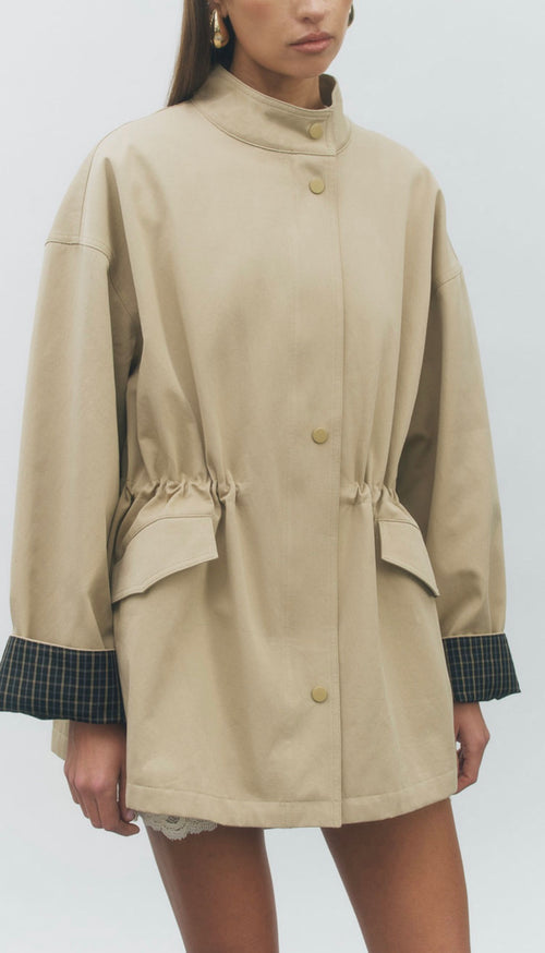 A model wears a beige oversized coat with a high stand collar, gold-tone snap buttons, a drawstring waist, and contrasting dark green plaid cuffs, shown against a light gray studio background.
