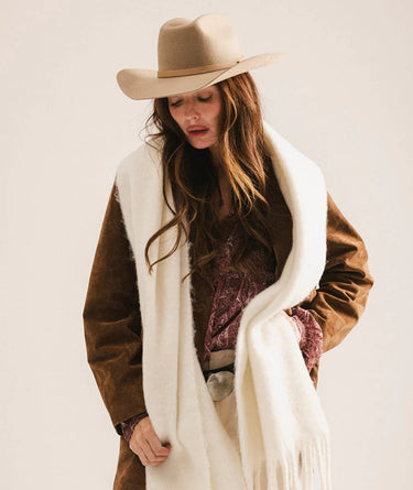 Woman wearing a brown coat, white scarf, and beige hat against a plain background