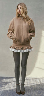 A woman modeling a brown cropped sweatshirt with pleated front detailing and a ruffled hem, paired with dark gray tights and pointed shoes, standing against a textured light gray wall.