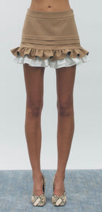 A model's lower body wearing a beige ruffled mini skirt with white ruffle trim and snakeskin-patterned high heels, standing against a light gray background.
