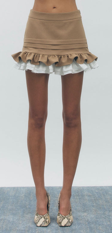 A model's lower body wearing a beige ruffled mini skirt with white ruffle trim and snakeskin-patterned high heels, standing against a light gray background.