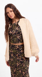A woman modeling a cream chunky knit cardigan over a black floral two-piece set, photographed against a plain white studio background.