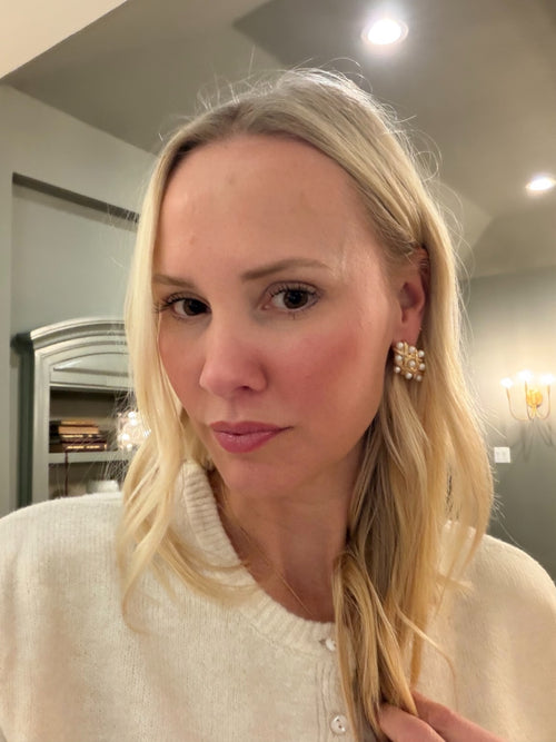 Woman with blonde hair wearing a white sweater and gold earrings indoors.